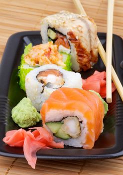 Arrangement of Various Maki Sushi with Salmon, Eel, Green Caviar and Crab on Black Plate with Wasabi, Marinated Ginger and Chopsticks on Straw mat background