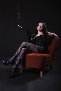 Young woman in retro dress with cigarette holder sitting on an old chair on dark background