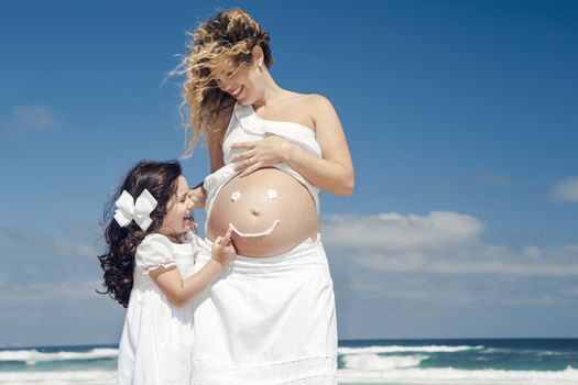 Beautiful pregnant woman in the beach with her little daugther making a smile on mom's belly with sunscreen