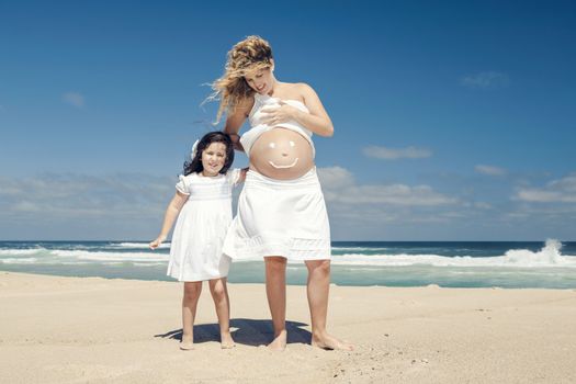 Beautiful pregnant woman in the beach with her little daugther making a smile on mom's belly with sunscreen