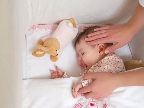 Hands of mother caressing her cute baby girl sleeping in a cot with pacifier and stuffed toy