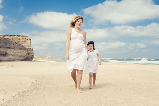 Beautiful pregnant woman and her little daughter walking on the beach