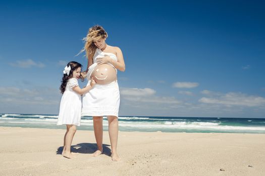 Beautiful pregnant woman in the beach with her little daugther making a smile on mom's belly with sunscreen