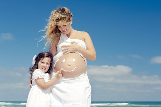 Beautiful pregnant woman in the beach with her little daugther making a smile on mom's belly with sunscreen