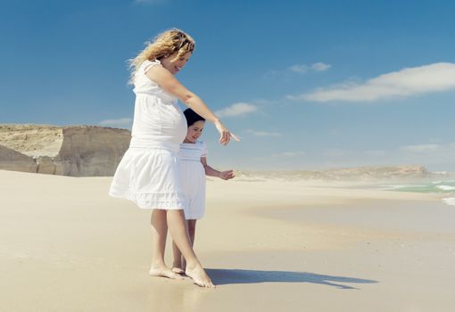 Beautiful pregnant woman and her little daughter on the beach pointing to the sand