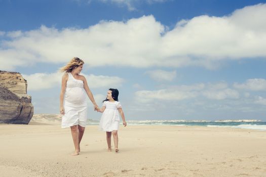 Beautiful pregnant woman and her little daughter walking on the beach