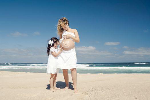 Beautiful pregnant woman in the beach with her little daugther making a smile on mom's belly with sunscreen