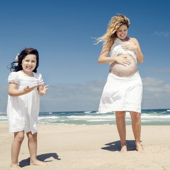 Beautiful pregnant woman in the beach with her little daughter applying sunscreen on mom's belly