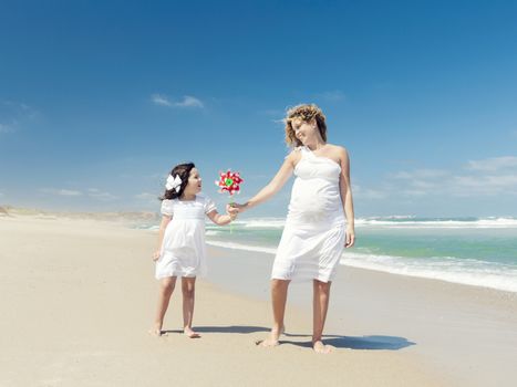 Beautiful pregnant woman and her little daughter really happy playing on the beach with a windmill
