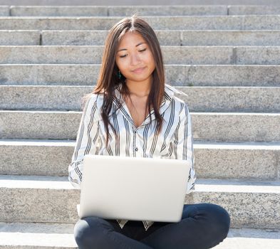 Smiling asian woman sitting on stairs and using a laptop