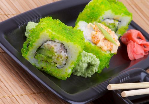Delicious Maki Sushi with Avocado and Green Caviar on Black Plate with Pair of Chopsticks on Straw mat background