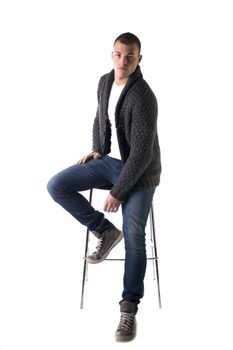 Attractive young man sitting on stool with wool sweater and jeans, isolated on white background