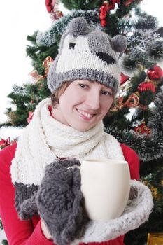 Smiling woman in knitted hat and mitten under Christmas tree with cup, focus on woman