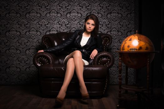 The beautiful young girl with dark hair in a black leather jacket and a white dress sits in a brown leather seat against a dark background