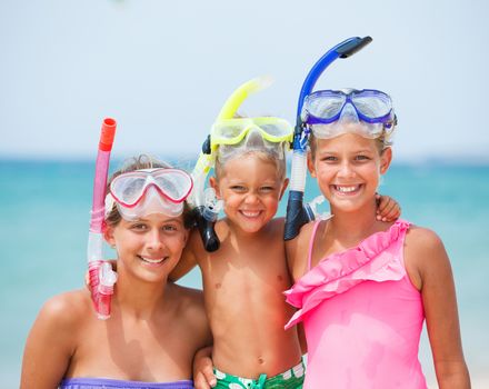 Closeup portrait of three happy children on beach with colorful face masks and snorkels, sea in background.