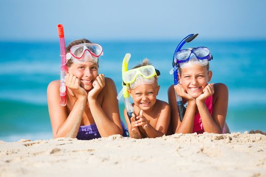 Three happy children on beach with colorful face masks and snorkels, sea in background.