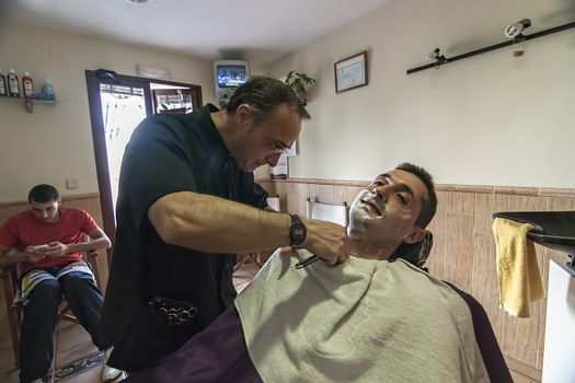 Barber shaving with a razor to young man in a barber's shop, Sabiote, Jaen province, Andalucia, Spain