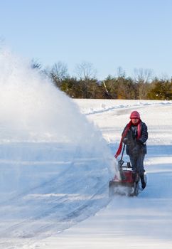 Senior lady using a snowblower on rural drive on windy day with a cloud or blizzard of snow blowing in the air