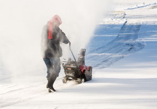 Senior lady using a snowblower on rural drive on windy day with a cloud or blizzard of snow blowing in the air