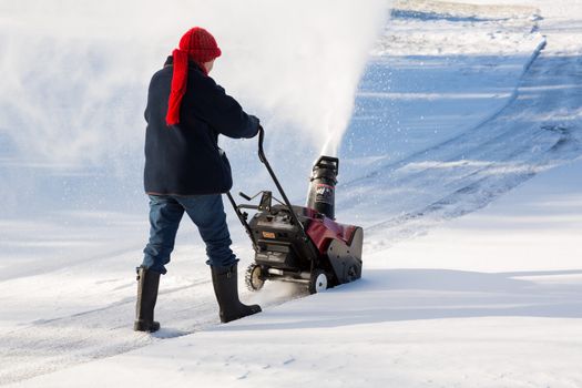 Senior lady using a snowblower on rural drive on windy day with a cloud or blizzard of snow blowing in the air