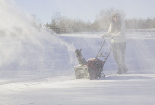 Young lady using a snowblower on rural drive on windy day with a cloud or blizzard of snow blowing in the air and covering her with icy snow