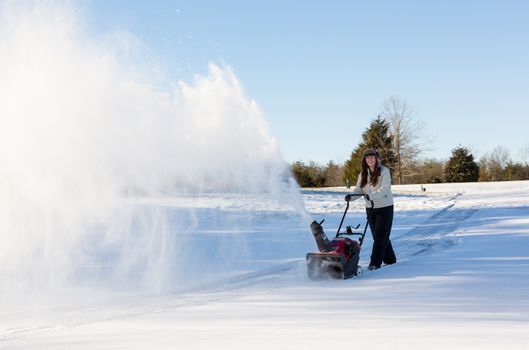 Young lady using a snowblower on rural drive on windy day with a cloud or blizzard of snow blowing in the air