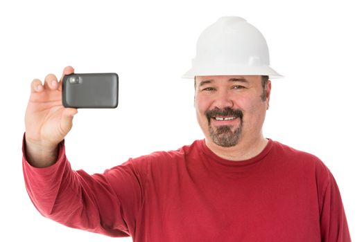 Smiling workman with a goatee beard wearing a hardhat taking a self-portrait using his smartphone, isolated on white
