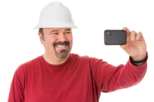 Workman with a neat goatee beard weaning a hardhat posing for a self-portrait giving a cheesy grin as he looks into the camera on his mobile phone, isolated on white