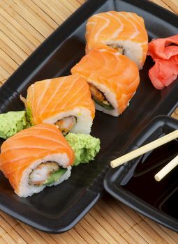 Arrangement of Philadelphia Sushi with Wasabi, Marinated Ginger and Soy Sauce in Black Square Shape Bowls with Pair of Chopsticks on Straw mat background