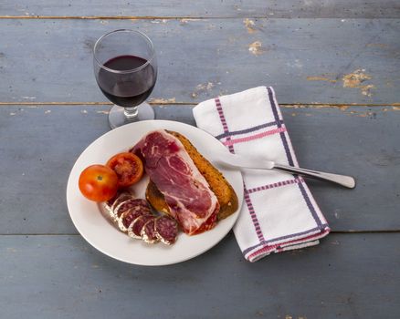 Still life with a catalan 'pa amb tomata' (bread or toast with tomato, salt and olive oil) with jam and cured meats, and a glass of red wine, on and old blue table