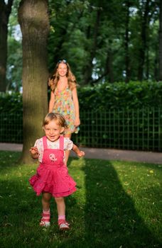 The smiling little girl run on the green lawn in the park with mother on the background. 