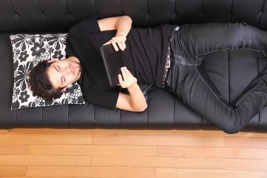 A young hispanic man lying on the sofa holding a Tablet PC.