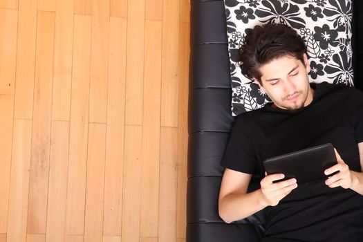 A young hispanic man lying on the sofa holding a Tablet PC.