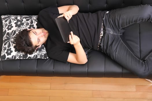 A young hispanic man lying on the sofa holding a Tablet PC.