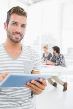 Portrait of a smiling young man using digital tablet with colleagues in background at a creative bright office