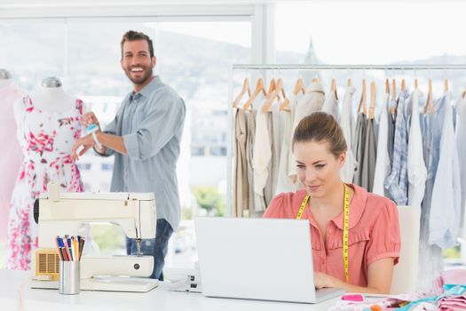 Young woman using laptop with male fashion designer working in background at the studio