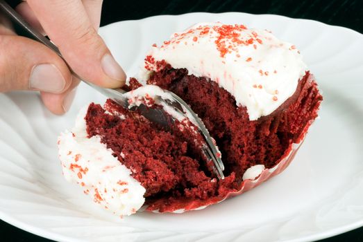 Close-up of a red velvet cupcake on a plate with a man's hand holding a fork slicing into it.