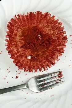Close-up of an empty red velvet cupcake wrapper lying on a plate covered in crumbs with a fork beside it.