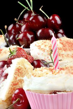 Close-up of a cream cheese iced strawberry cherry muffin with a pink candle surrounded by fruit and other muffins, topped with graham cracker crumbs. Vertical.