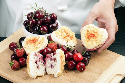 Close-up of a chef displaying a cream cheese iced strawberry cherry muffin surrounded by fruit and other muffins, topped with graham cracker crumbs.