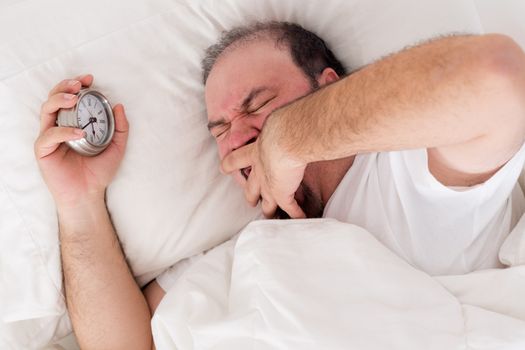 Man lying in bed yawning as he tries to wake up with his alarm clock clutched in his hand