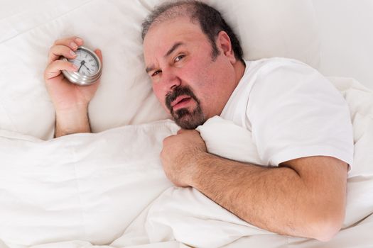 Irascible man glaring balefully at the camera as he lies in bed holding the alarm clock in his hand after a sleepless night wanting to remain in bed until noon