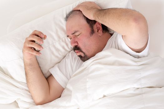Man suffering from a hangover clutching his head in one hand and alarm clock in the other as he suffers in the morning wondering what happened