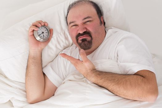 Middle-aged bearded man kept awake by noisy neighbours lying in bed grimacing and pointing to the time on his alarm clock