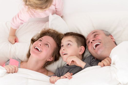 Playful little girl with her family in bed clambering over the top of the pillow making them laugh with amusement and pleasure as her brother and parents snuggle together under the bedclothes