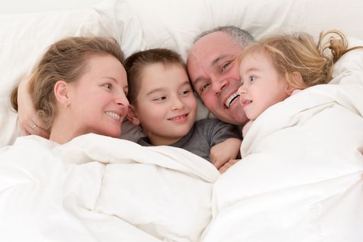 Happy young family cuddling together in bed with a pretty young girl and her brother enjoying the love and devotion of their parents as they smile and look at each other, close up of their expressions