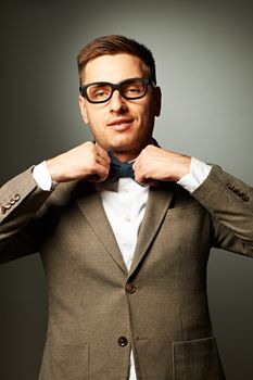 Confident nerd in eyeglasses adjusting his bow-tie against grey background