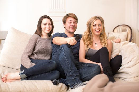 Three attractive young teenage friends sitting close together on a comfortable sofa watching television and smiling in appreciation of the program