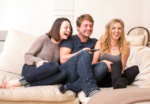 Three teenagers enjoying a funny television program laughing merrily as they sit and relax together on a comfortable sofa, one boy and two girls