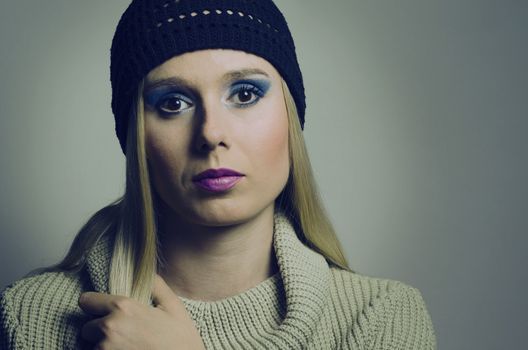 fashion portrait of a blonde woman in studio wearing a heavy make up and a turtleneck sweater with a black knitted hat